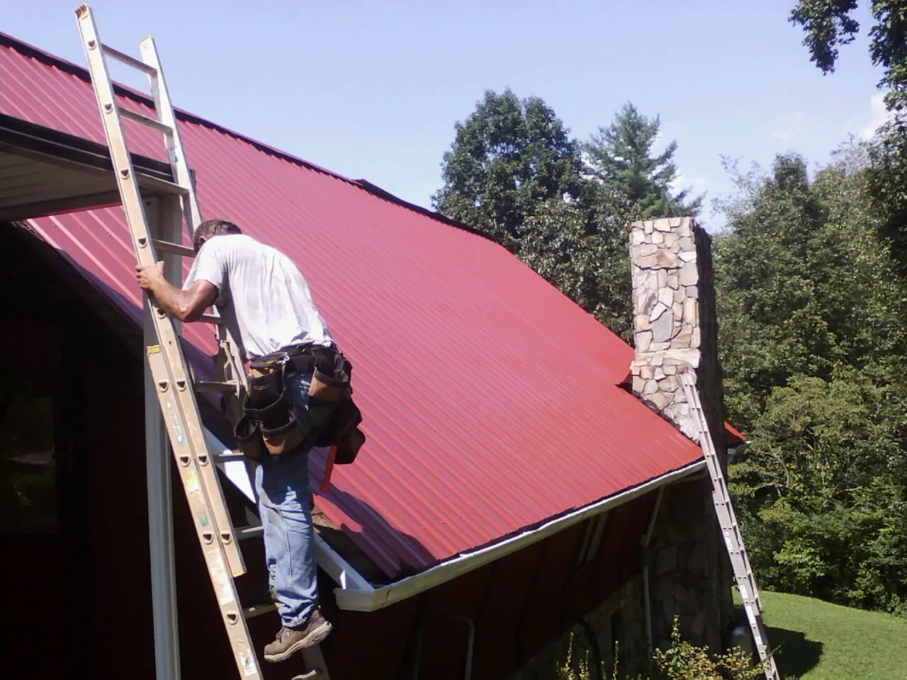 Roofer on a ladder beside a red metal roof and stone chimney — Soffit & Fascia Repair by Roofing Brothers in Cathlamet